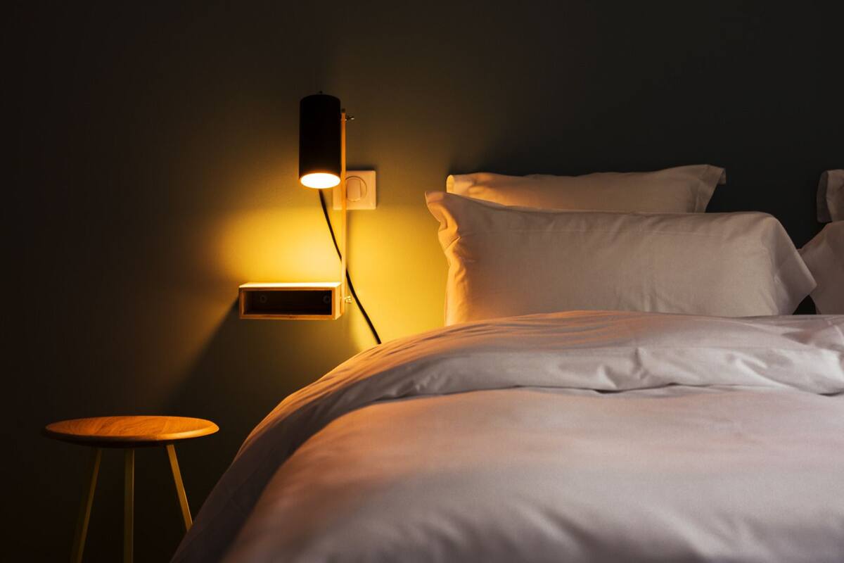 Un lit bien fait, avec du linge de lit blanc, une table de chevet et un luminaire fixé au mur.