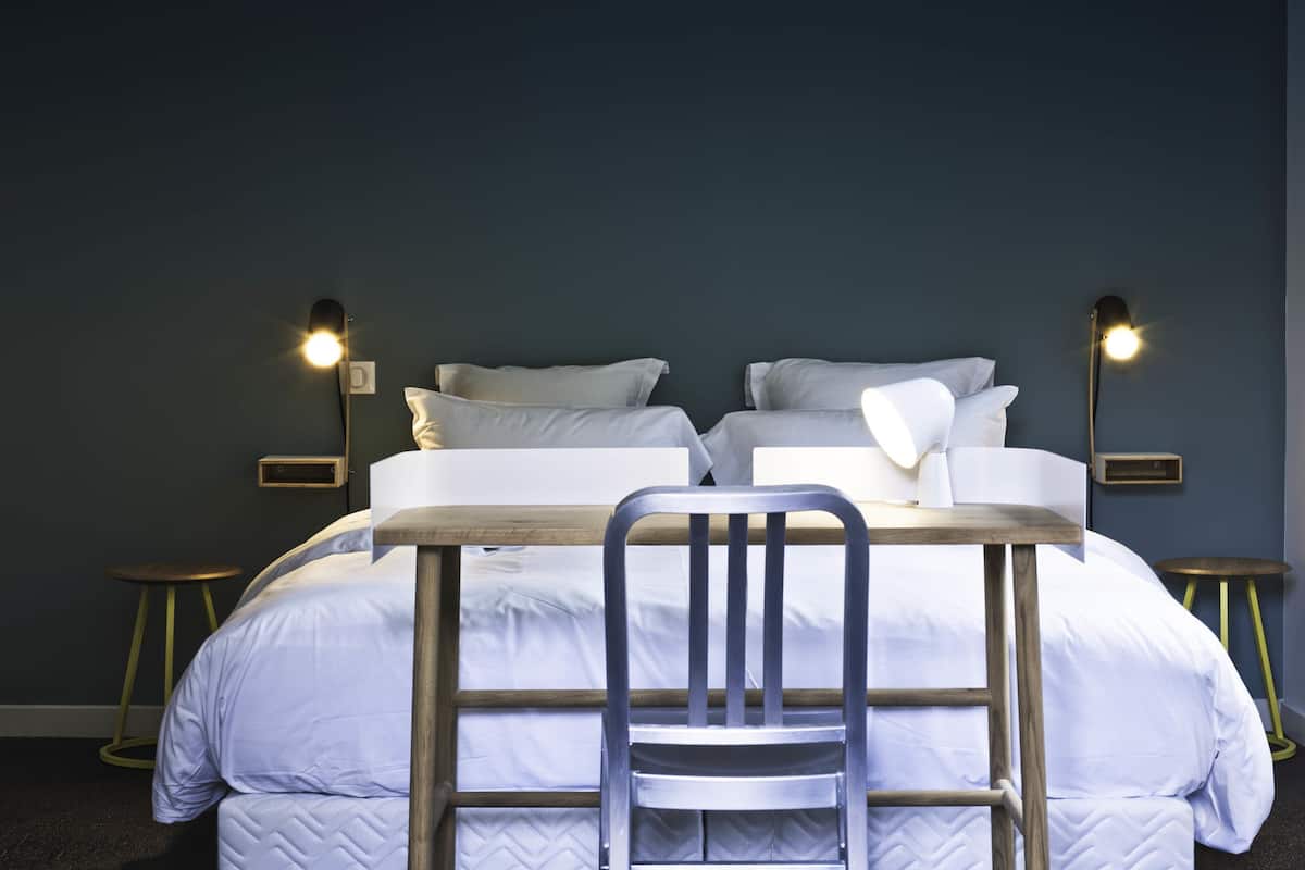Un lit bien fait, avec du linge de lit blanc, deux lampes de chevet, une petite table en bois et une chaise placée devant le lit.