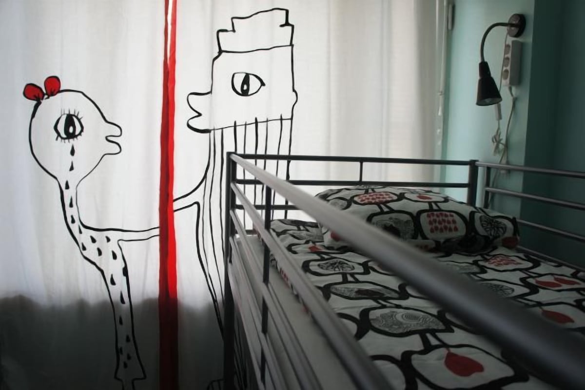 A child's drawing on a curtain, a bunk bed with patterned bedding, and a wall-mounted lamp.