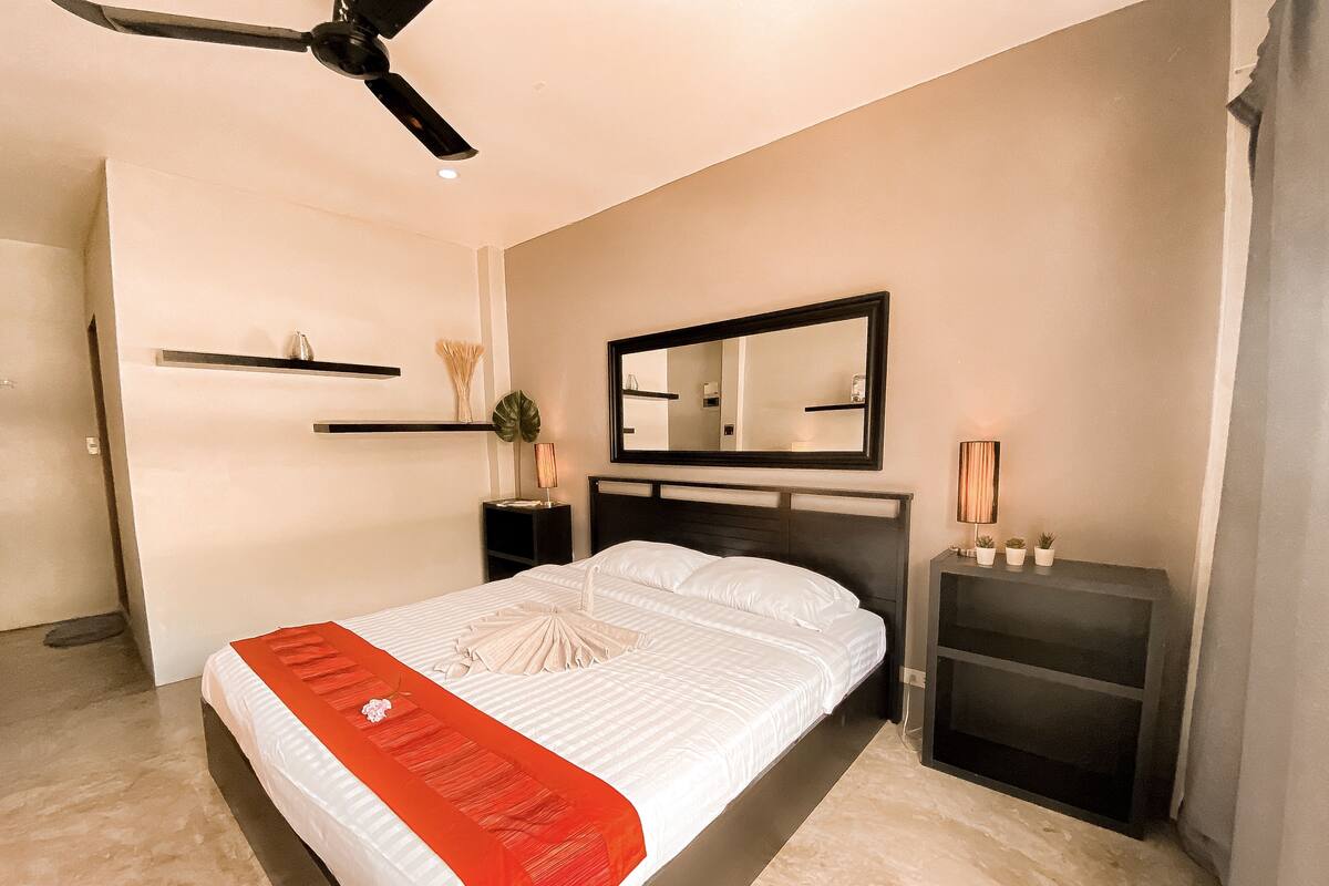 A modern bedroom with a bed, a ceiling fan, a nightstand, a mirror, and a small plant.