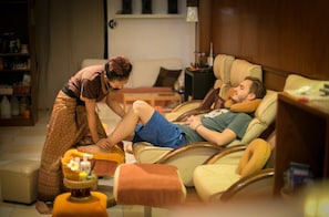 A man lying on a beige sofa in a living room with a wooden floor, a small round table with a bottle and a flower, and a bookshelf in the background.