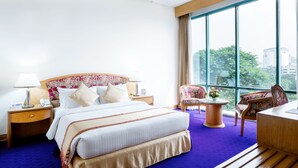 A hotel room with a large bed, a desk, a chair, and a view of the city through large windows. - Hotel Lake Castle (Dhaka)