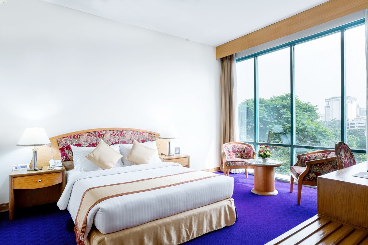 Een hotelkamer met een groot bed, een bureau, een stoel, een nachtkastje en uitzicht op de stad door grote ramen.