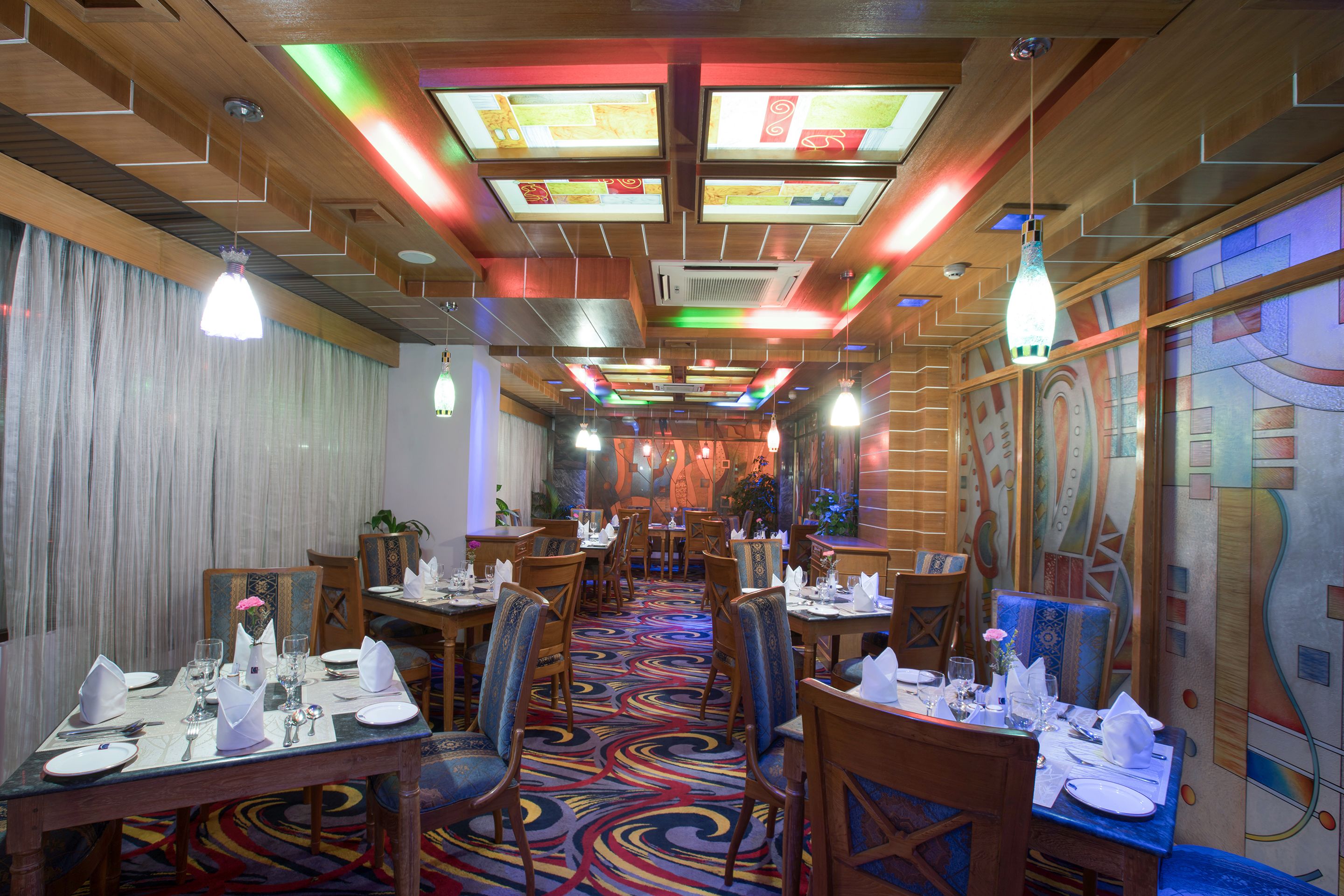 A restaurant with wooden ceiling panels, colorful carpet, and tables set for dining.