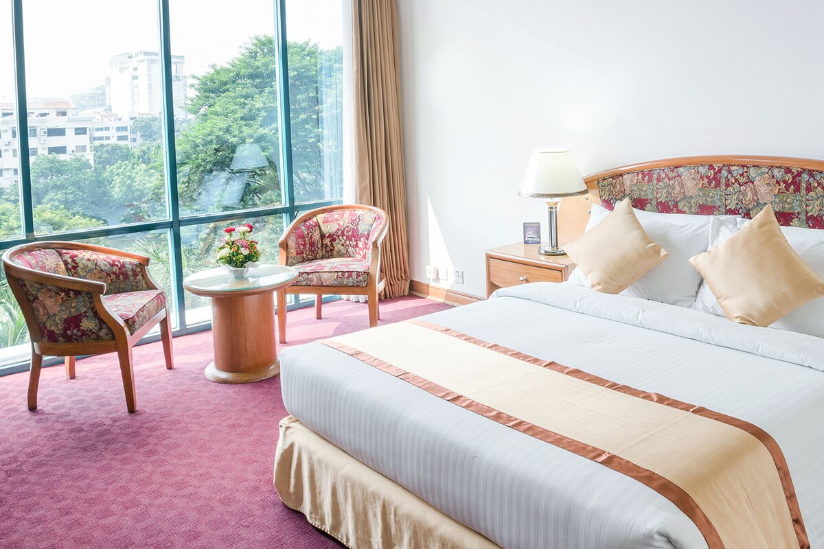 A hotel room with a large bed, two chairs, a small table, and a view of buildings and trees through large windows.