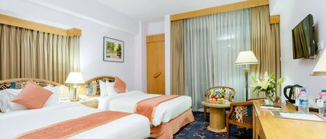 A hotel room with two beds, a desk, a chair, a TV, a lamp, and a small table with a flower vase.