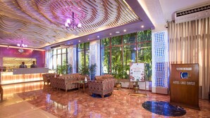 A hotel lobby with a reception desk, patterned sofas, a large window with a view of greenery, and a concierge desk. - Hotel Lake Castle (Dhaka)