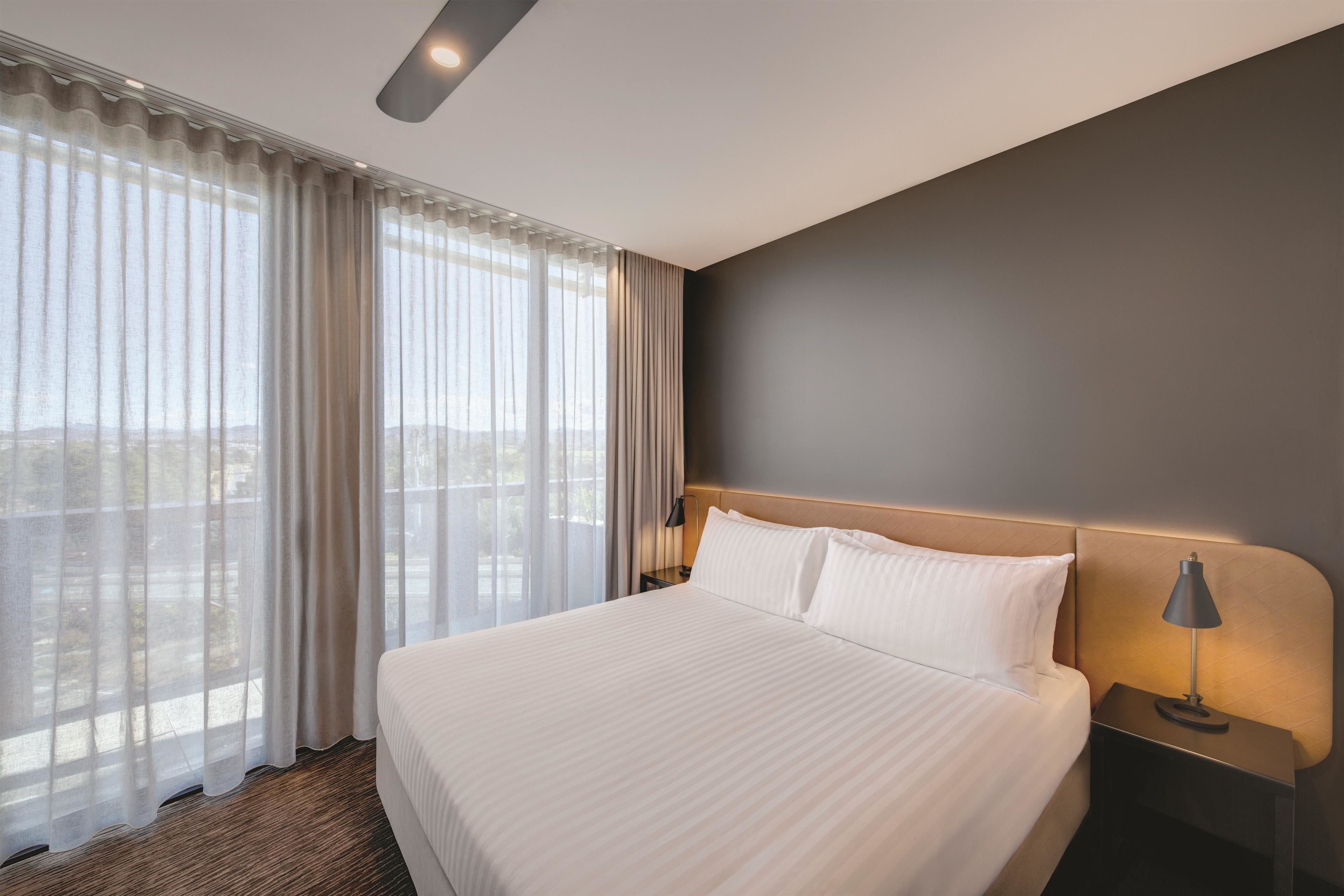 A hotel room with a large bed, a nightstand, a lamp, and a view of the city through sheer curtains.