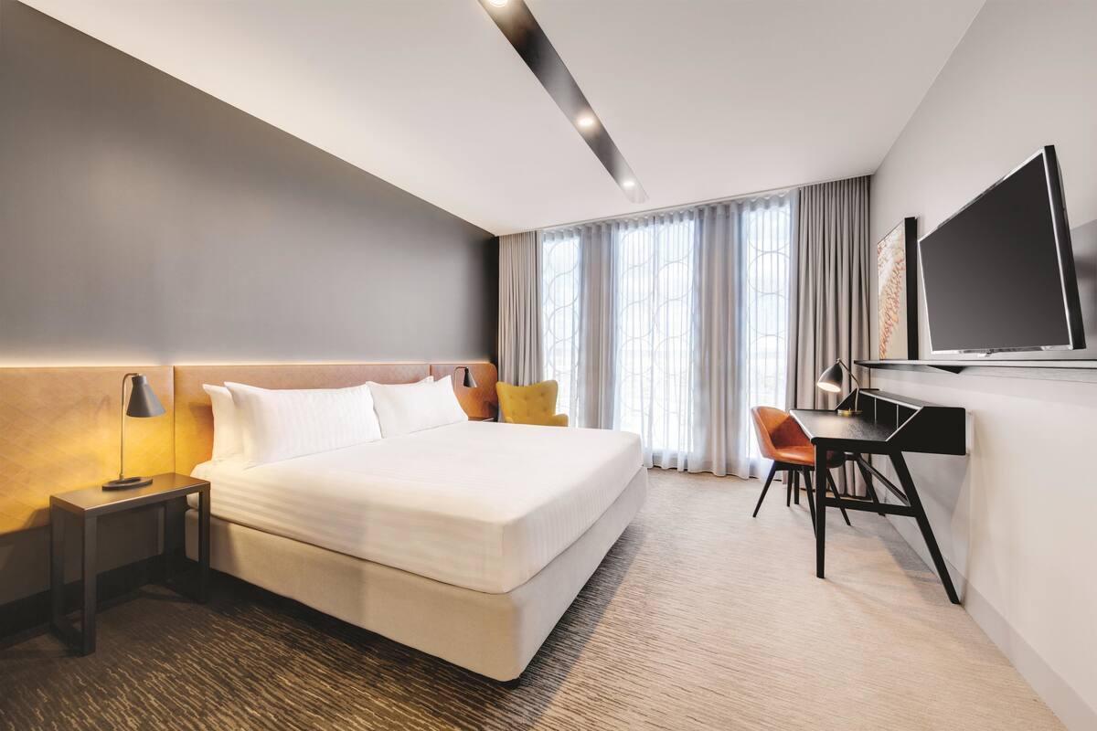 A modern hotel room with a large bed, a desk, a TV, and a window with curtains.