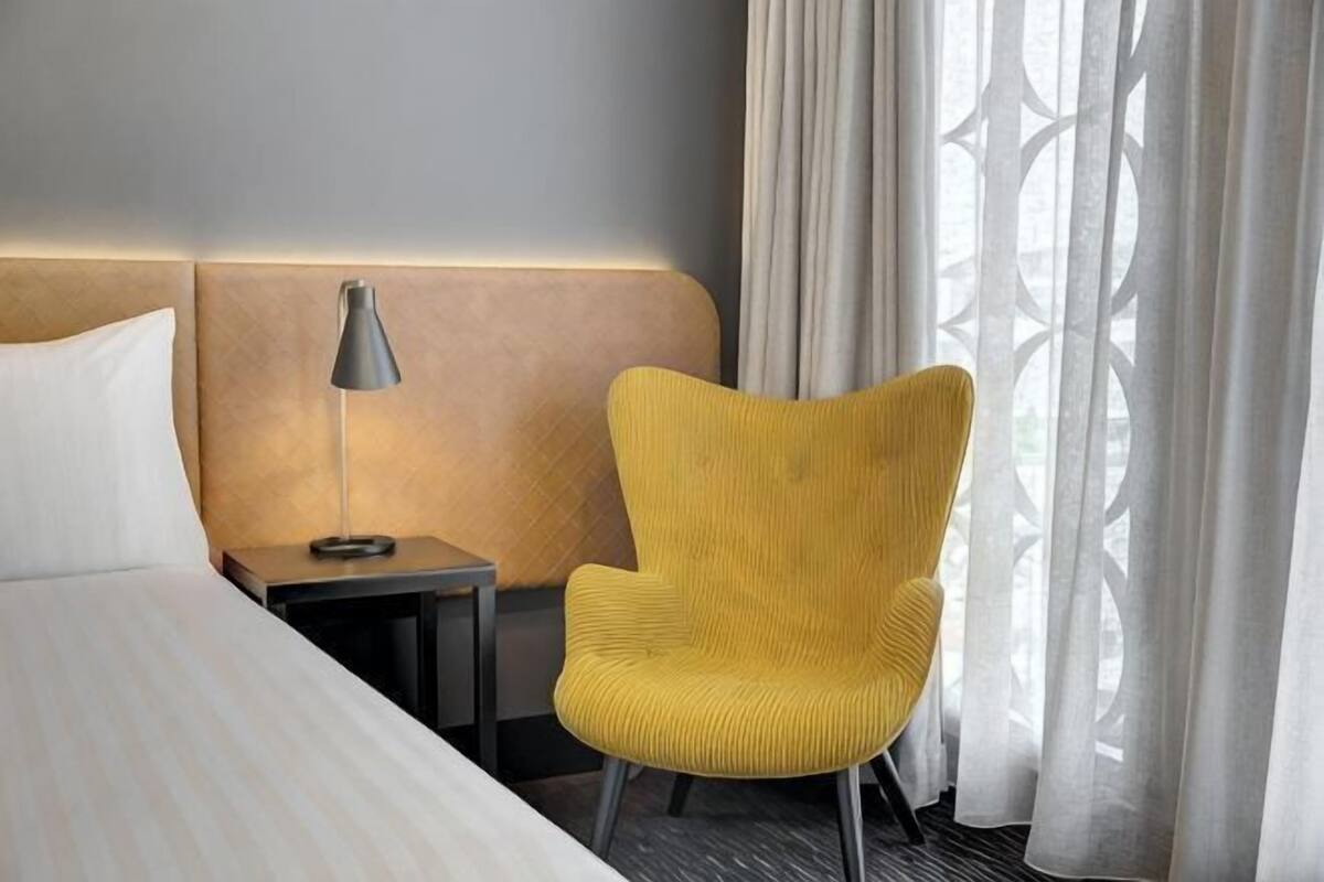 A hotel room with a bed, a nightstand with a lamp, a yellow chair, and a window with curtains.