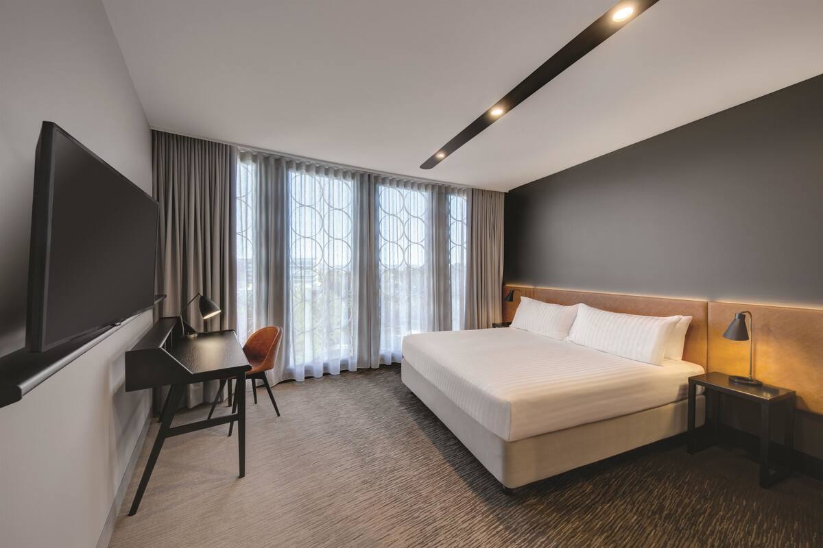 A modern hotel room with a large bed, a desk with a chair, a TV, and a nightstand with a lamp.