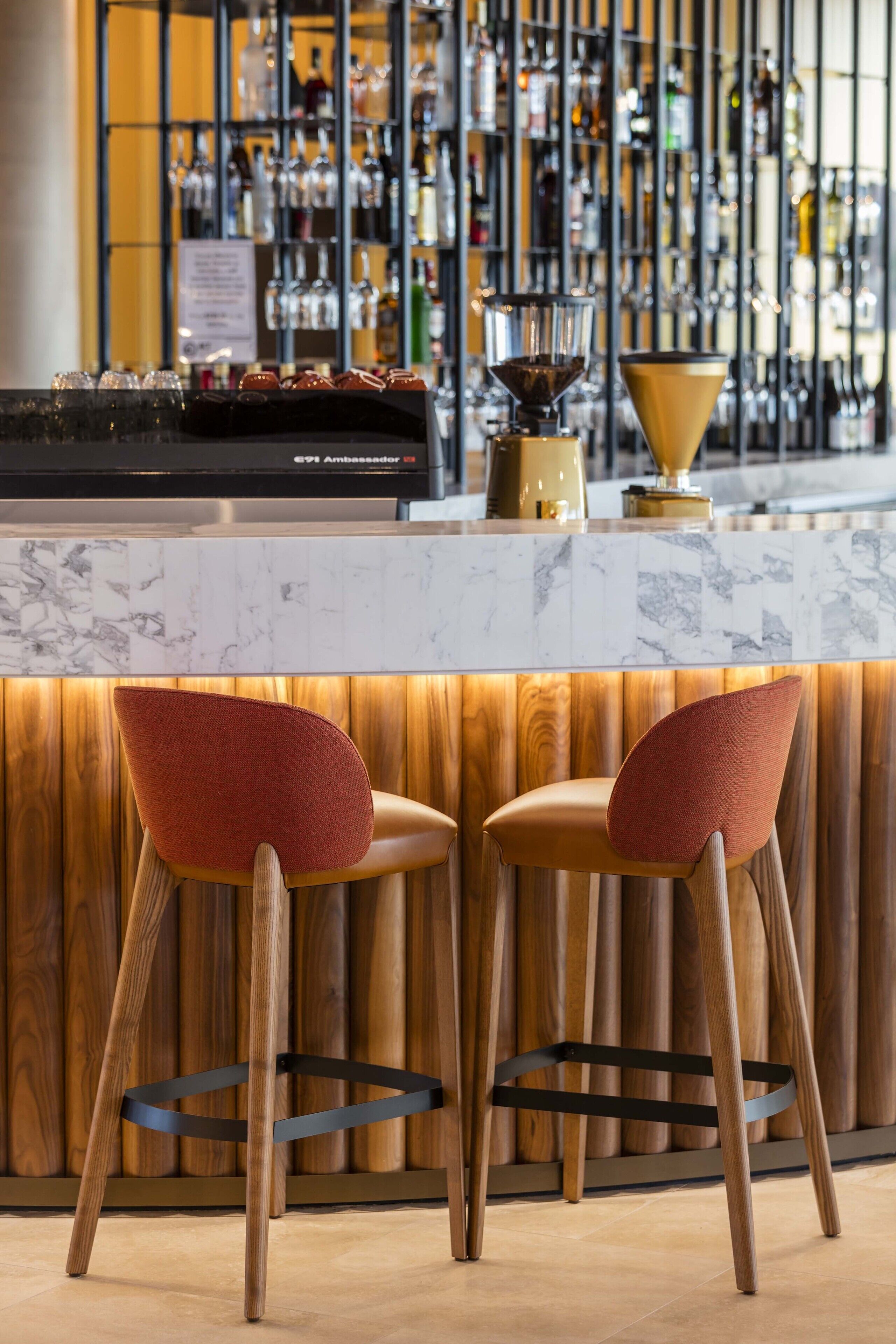 A bar area with a marble countertop, two red upholstered bar stools, and a variety of bottles and glasses in the background.