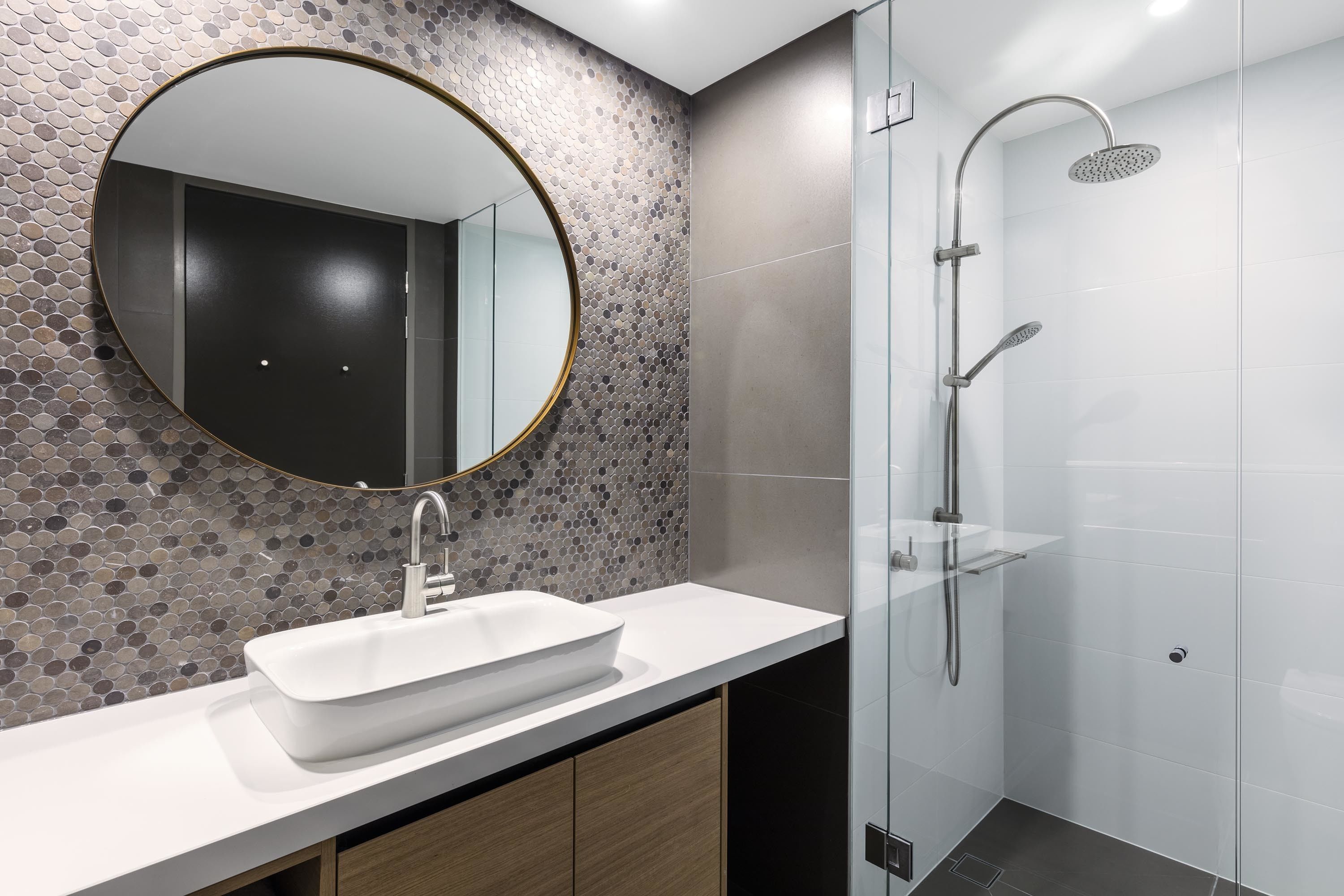 Un baño moderno con un espejo redondo, un lavamanos blanco y una zona de ducha con mampara de vidrio.
