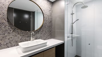 Un baño moderno con un espejo redondo, un lavamanos blanco y una zona de ducha con mampara de vidrio.