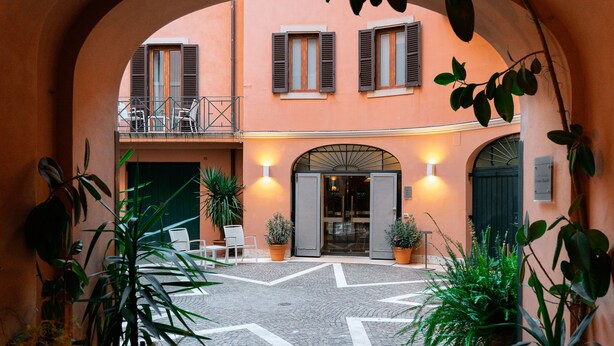 An arched courtyard with a cobblestone floor, potted plants, and a building with a balcony and shutters. - Unica Suites Rome (Rome)