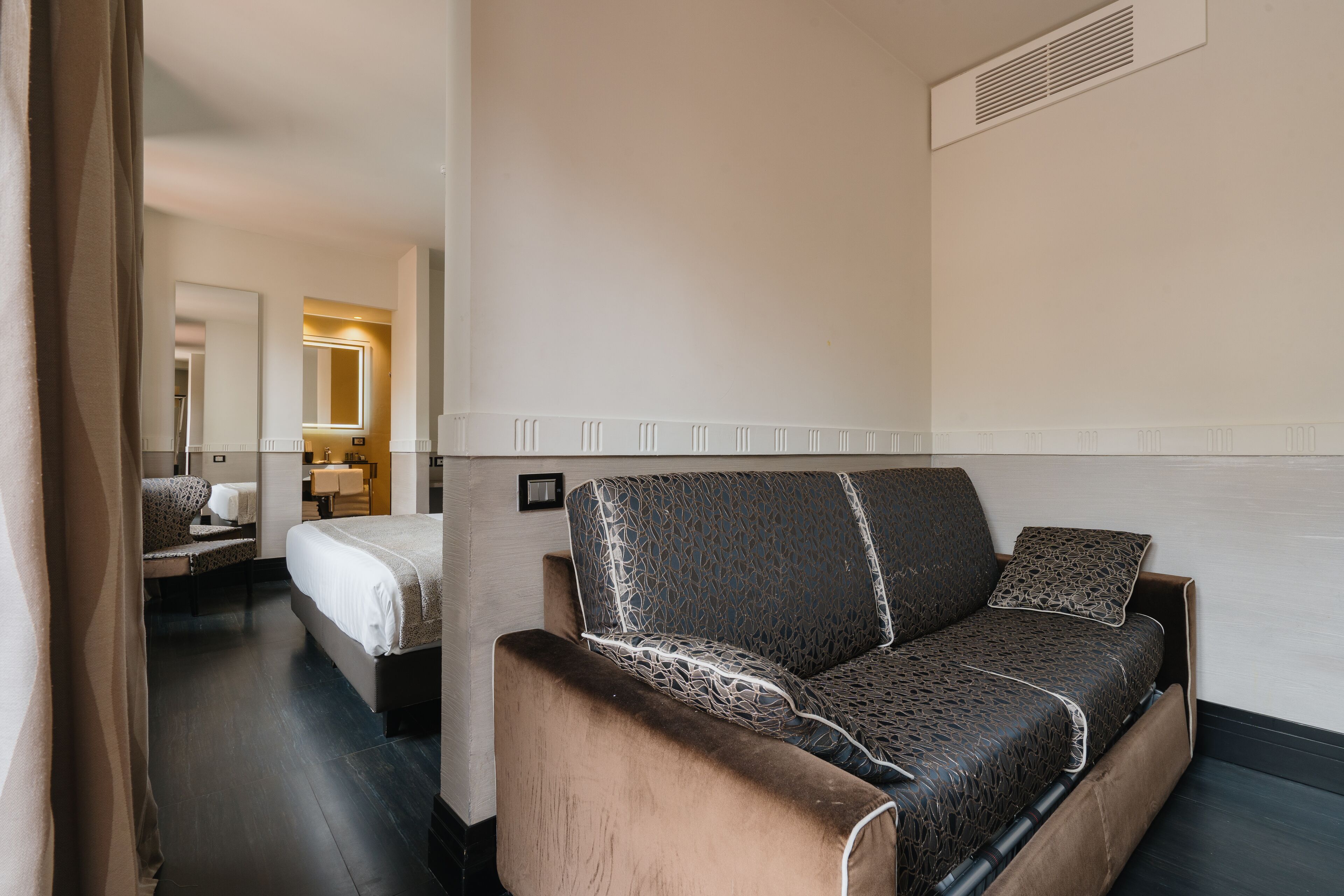 A hotel room with a sofa, a bed, a chair, and a mirror.
