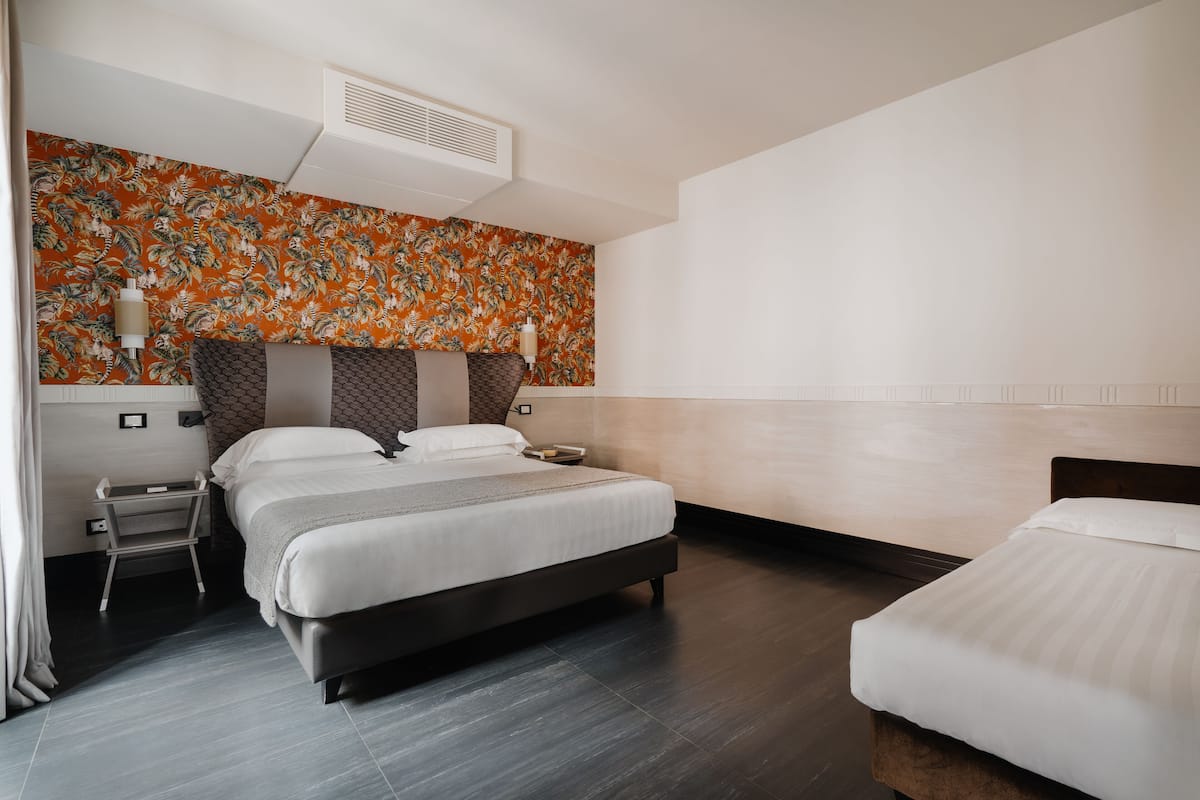A modern hotel room with a large bed, a smaller bed, a nightstand, a wall-mounted air conditioner, and a decorative wall with a tropical pattern.