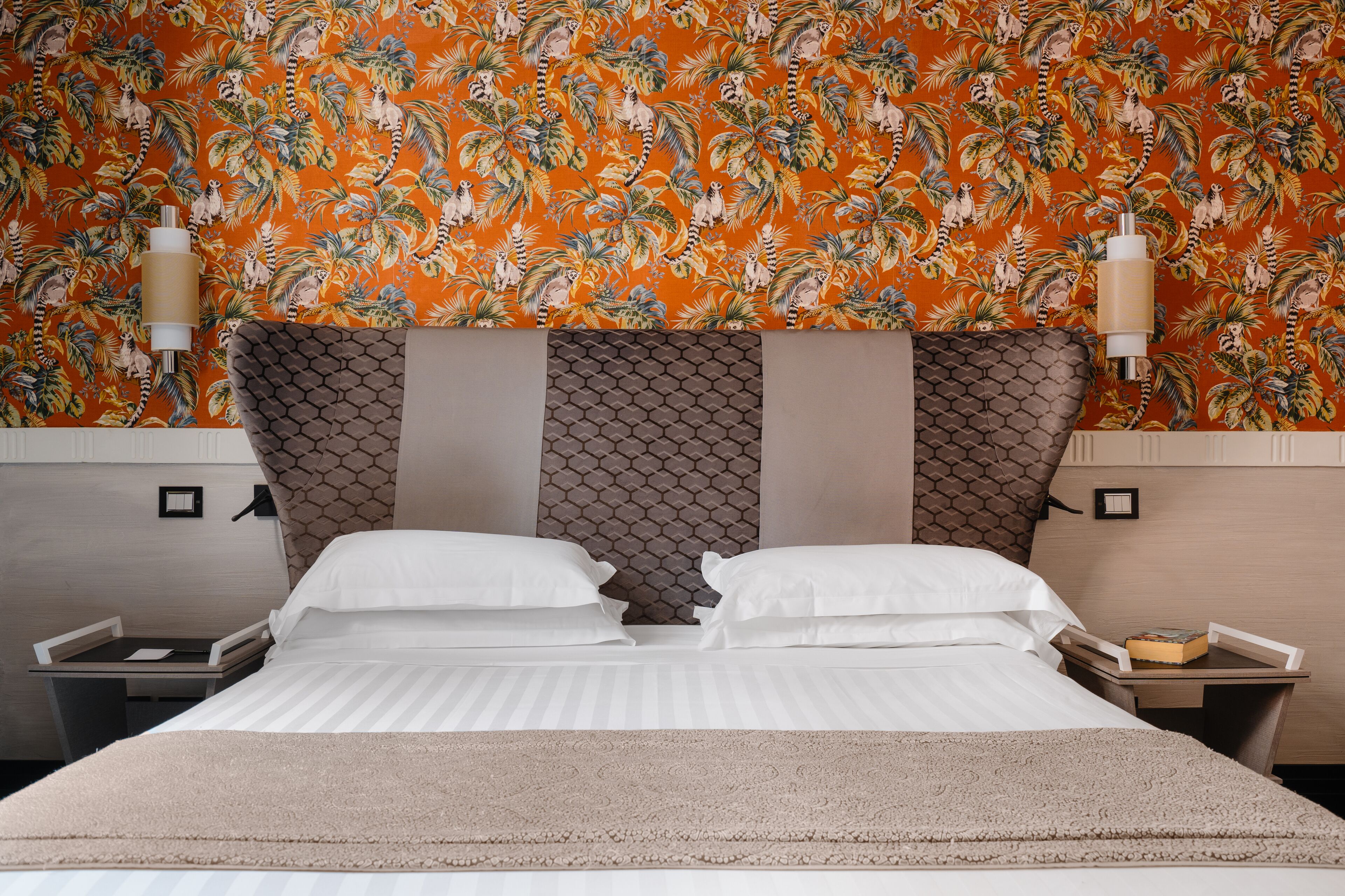 A hotel room with a bed, two bedside tables, a nightstand, a decorative headboard, and a wall with a tropical pattern.