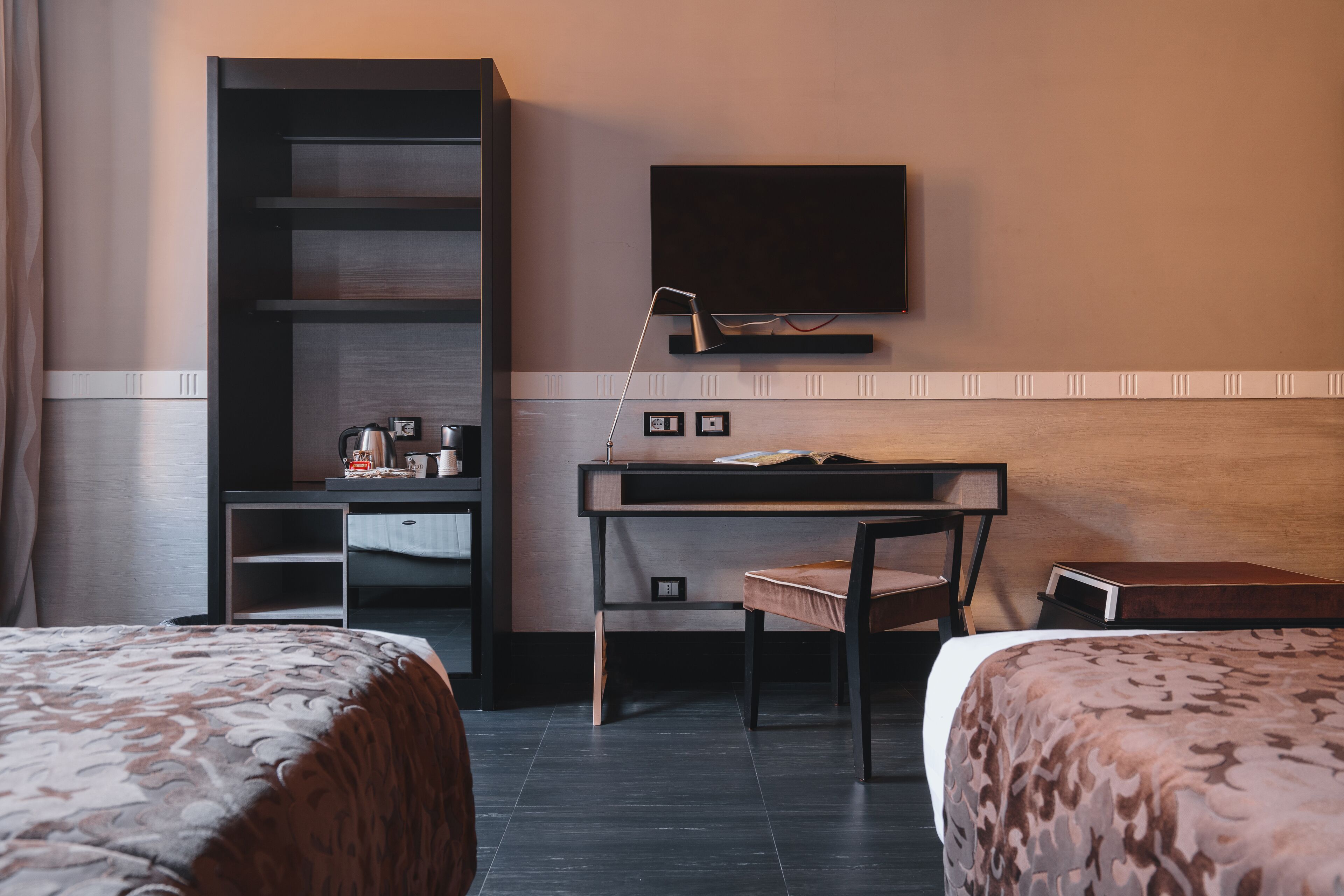 A hotel room with two beds, a desk with a chair, a television mounted on the wall, and a bookshelf.