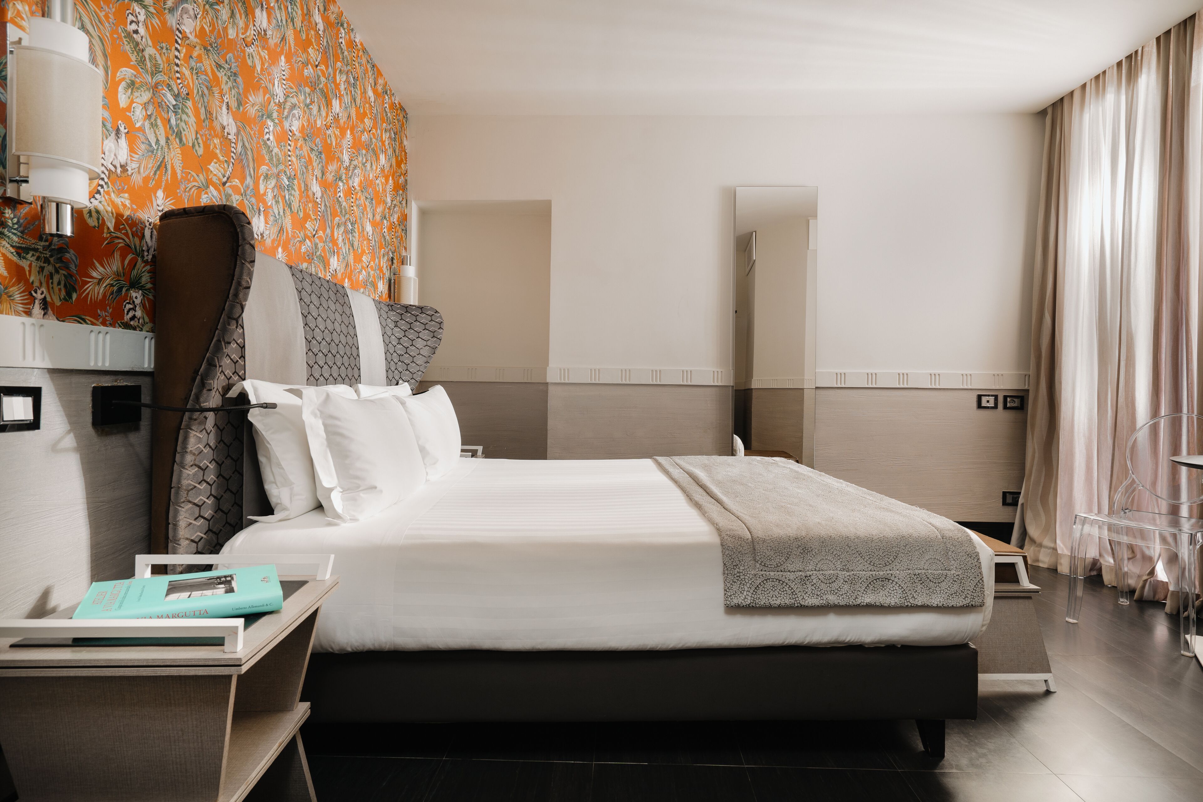 Une chambre d’hôtel moderne avec un grand lit, une table de chevet avec une lampe, un miroir et une vue sur l’océan par la fenêtre.