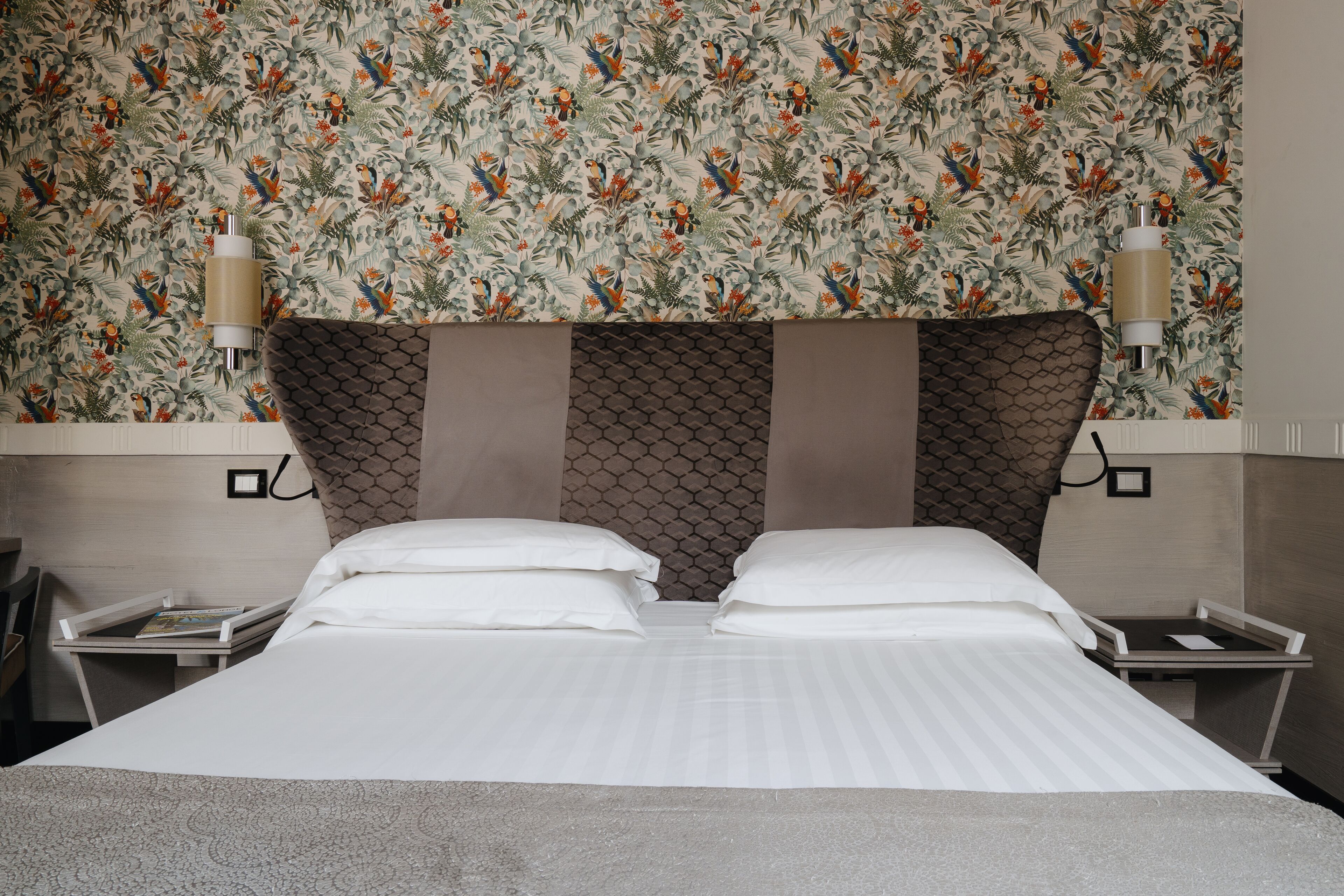 A hotel room with a bed, two bedside tables, and a wall with a floral pattern.