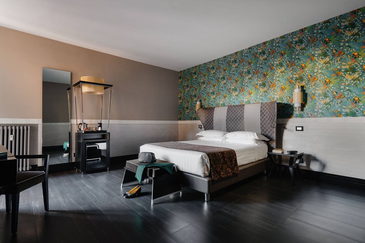 A modern hotel room with a large bed, a small desk, a chair, a nightstand, and a wall-mounted lamp.