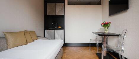 A compact hotel room with a bed, a small kitchenette, a dining area with a table and chairs, and a mounted TV.