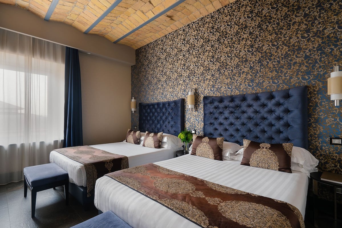 A hotel room with two beds, a large window with sheer curtains, a patterned wall, and a chandelier.