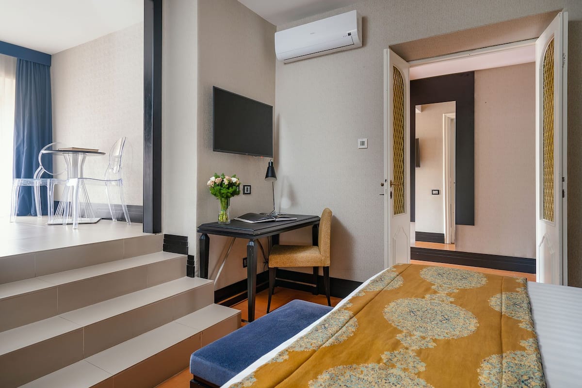 A modern hotel room with a bed, a desk with a chair, a TV mounted on the wall, and a staircase.