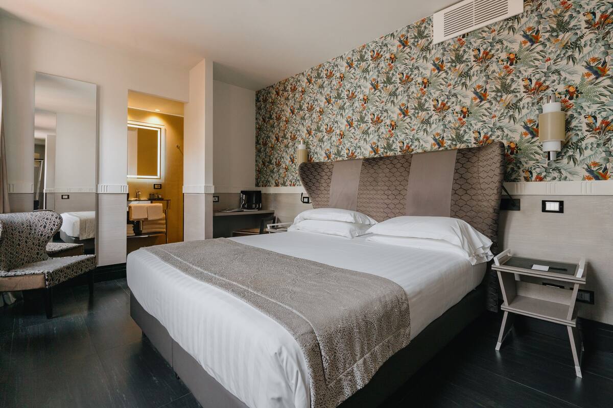 A hotel room with a large bed, a bedside table, a chair, and a mirror.