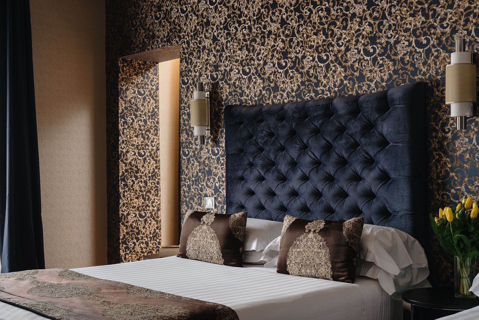 A bedroom with a large tufted headboard, a bed with multiple pillows, a nightstand with a lamp, and a vase with flowers.