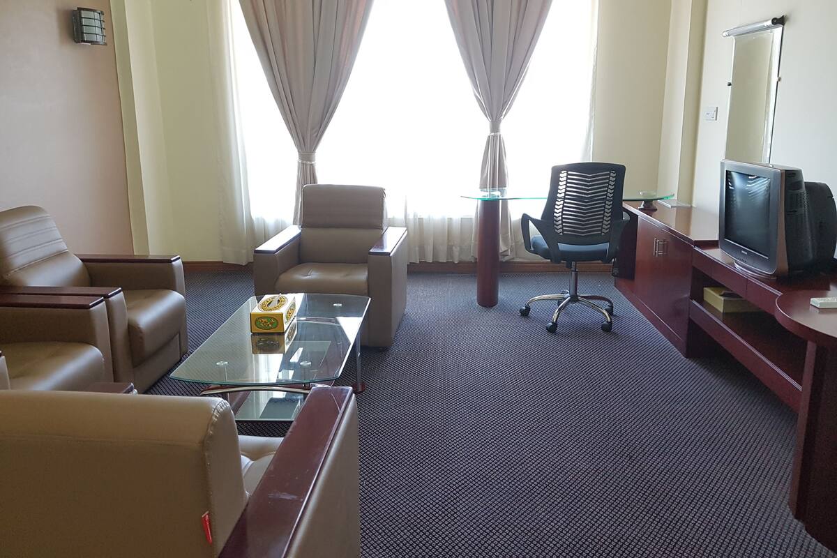 Une chambre d’hôtel avec un canapé, un fauteuil et une petite table sur laquelle se trouve une boîte. Il y a une télévision et une chaise près de la fenêtre.