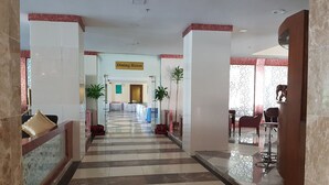 Un couloir d’hôtel avec un sol en marbre, une salle à manger visible au loin et des plantes en pot disposées le long des murs.