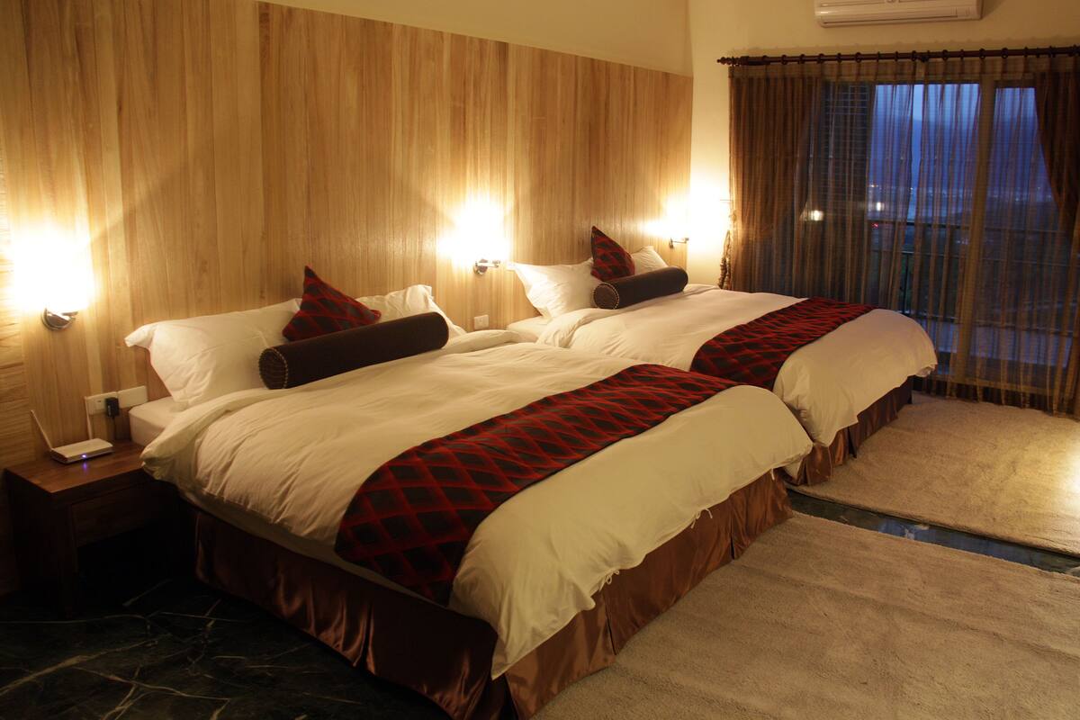 Une chambre d’hôtel avec deux lits, une tête de lit en bois, des lampes de chevet et une vue sur la ville, visible à travers les rideaux.