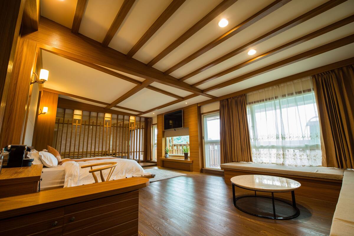 Une chambre d’hôtel avec un plafond en bois, un lit avec du linge de maison blanc, une petite table, un canapé et un téléviseur fixé au mur.