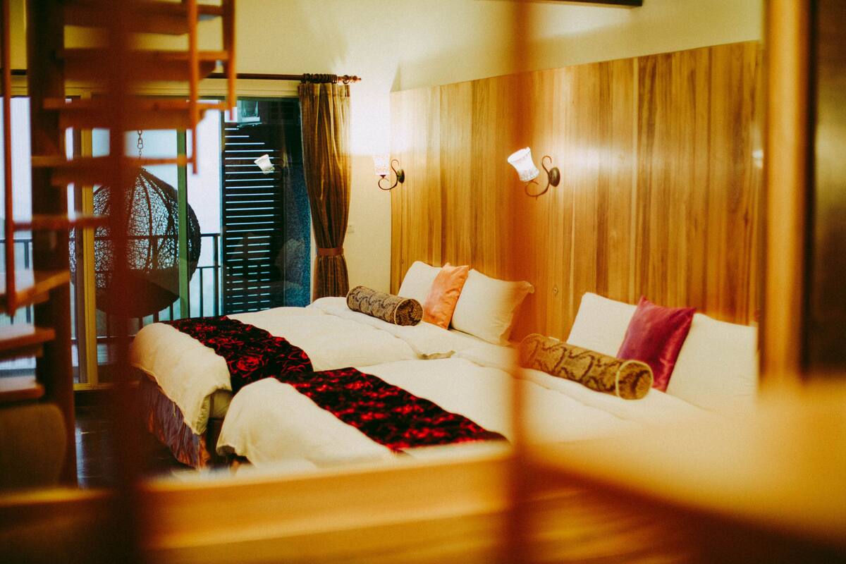 Une chambre d’hôtel avec une tête de lit en bois, deux lits, une grande fenêtre avec des stores et un balcon avec une chaise.
