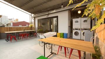 A rooftop terrace with a long wooden table, red stools, a bicycle, and a washing machine.