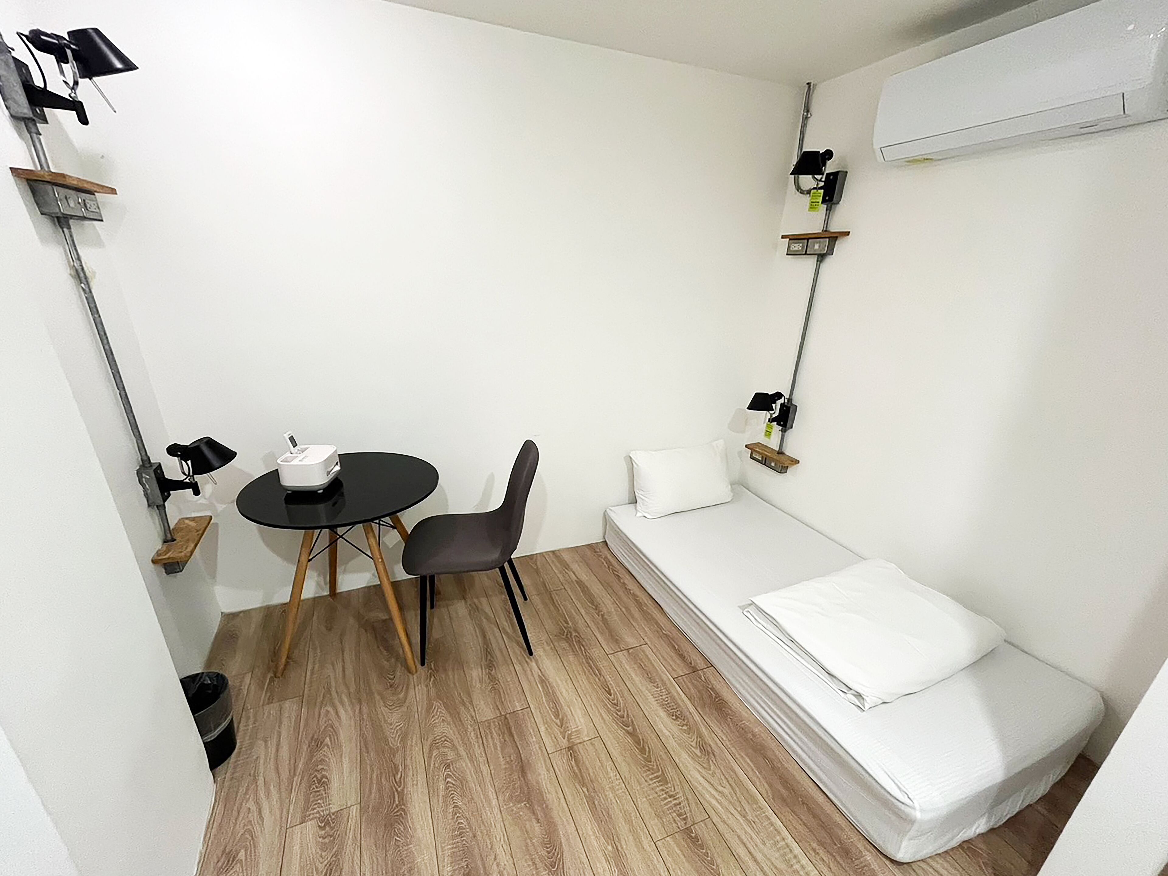 Una habitación pequeña y funcional con una cama individual, una mesita con una silla y una estantería con una lámpara.