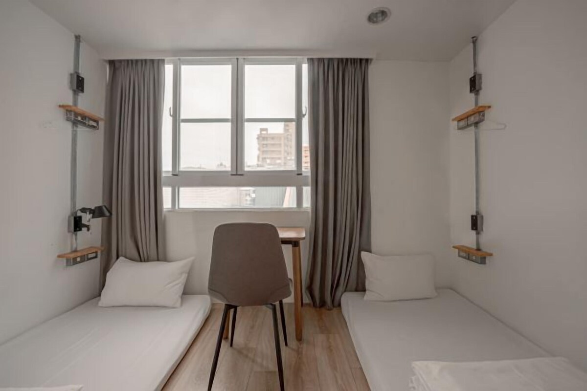 Une petite chambre moderne équipée de deux lits simples, d’un bureau et d’une chaise.
