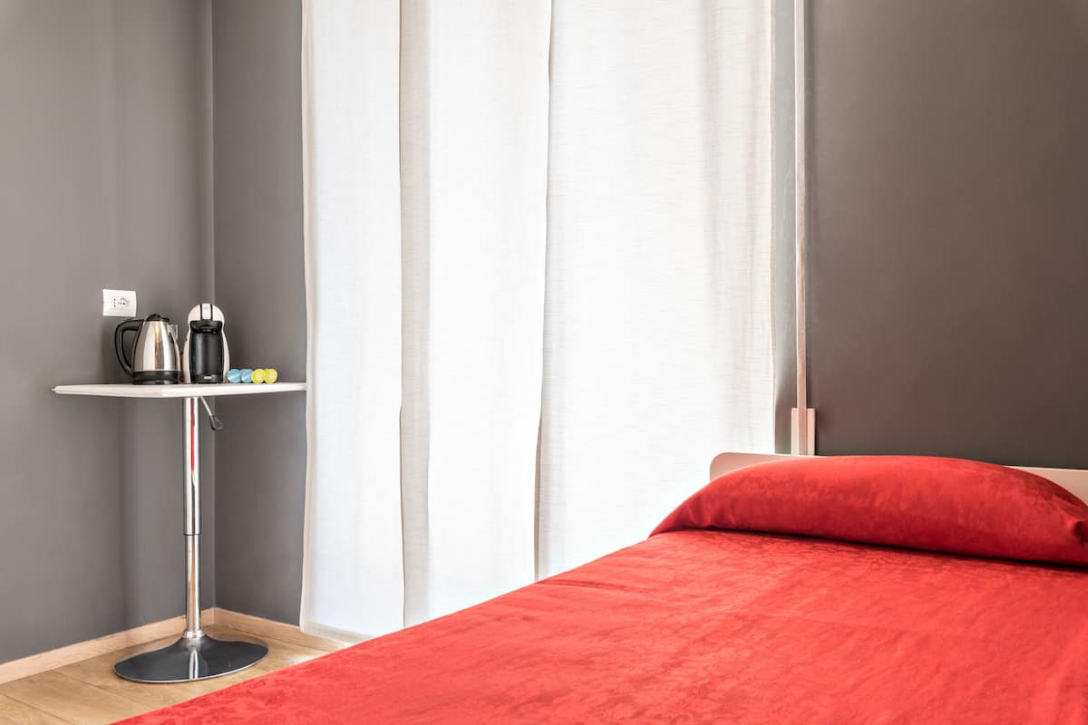 Une chambre avec un lit, une couverture rouge, une petite table avec une bouilloire et une fenêtre avec des rideaux.