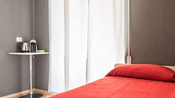 Une chambre avec un lit, une couverture rouge, une petite table avec une bouilloire et une fenêtre avec des rideaux.