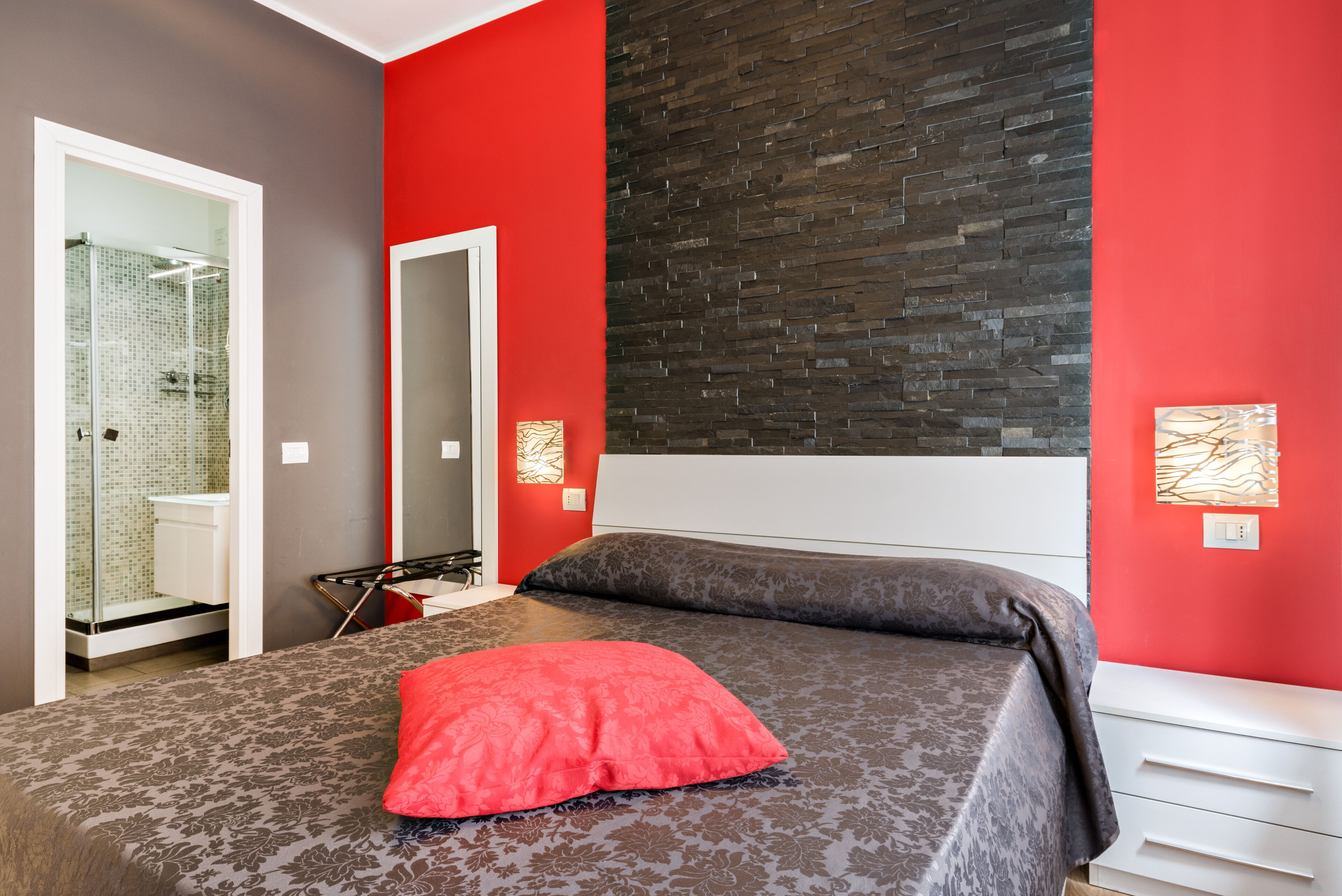 A bedroom with a bed, a red wall, a mirror, and a bathroom with a glass shower.