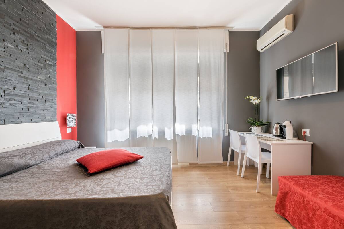 Une chambre d’hôtel moderne avec un lit gris, un oreiller rouge, un bureau avec une chaise, une plante en pot et un téléviseur à écran plat fixé au mur.
