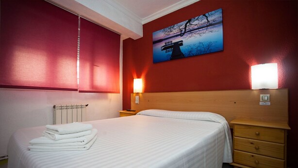 Habitación de hotel con cama, mesita de noche, lámpara de pared y un cuadro de un atardecer sobre un puente.