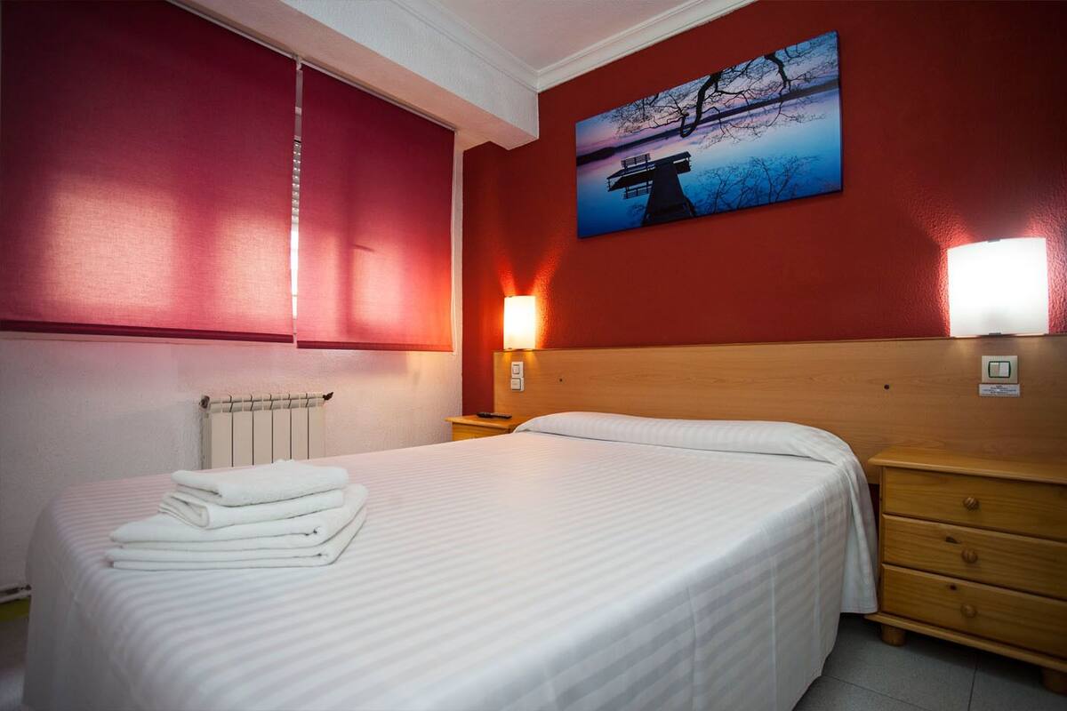 A hotel room with a bed, a nightstand, a wall-mounted lamp, and a painting of a sunset over a bridge.