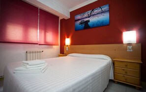 Une chambre d’hôtel avec un lit, une table de chevet, une lampe fixée au mur et un tableau représentant un coucher de soleil sur un pont.