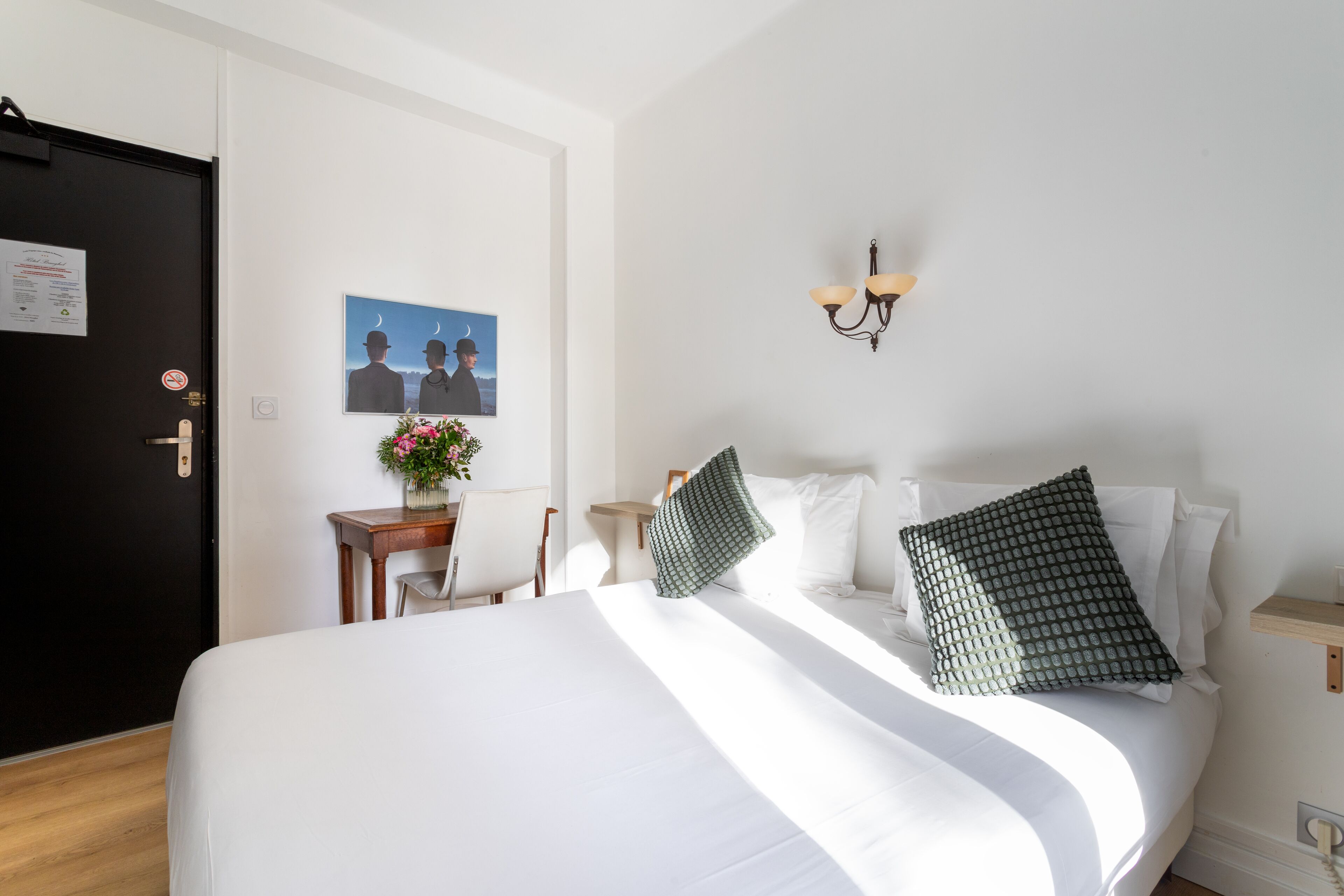Habitación de hotel con una cama grande, un escritorio con silla, una mesita con un florero y una lámpara de pared, además de un cuadro en la pared.