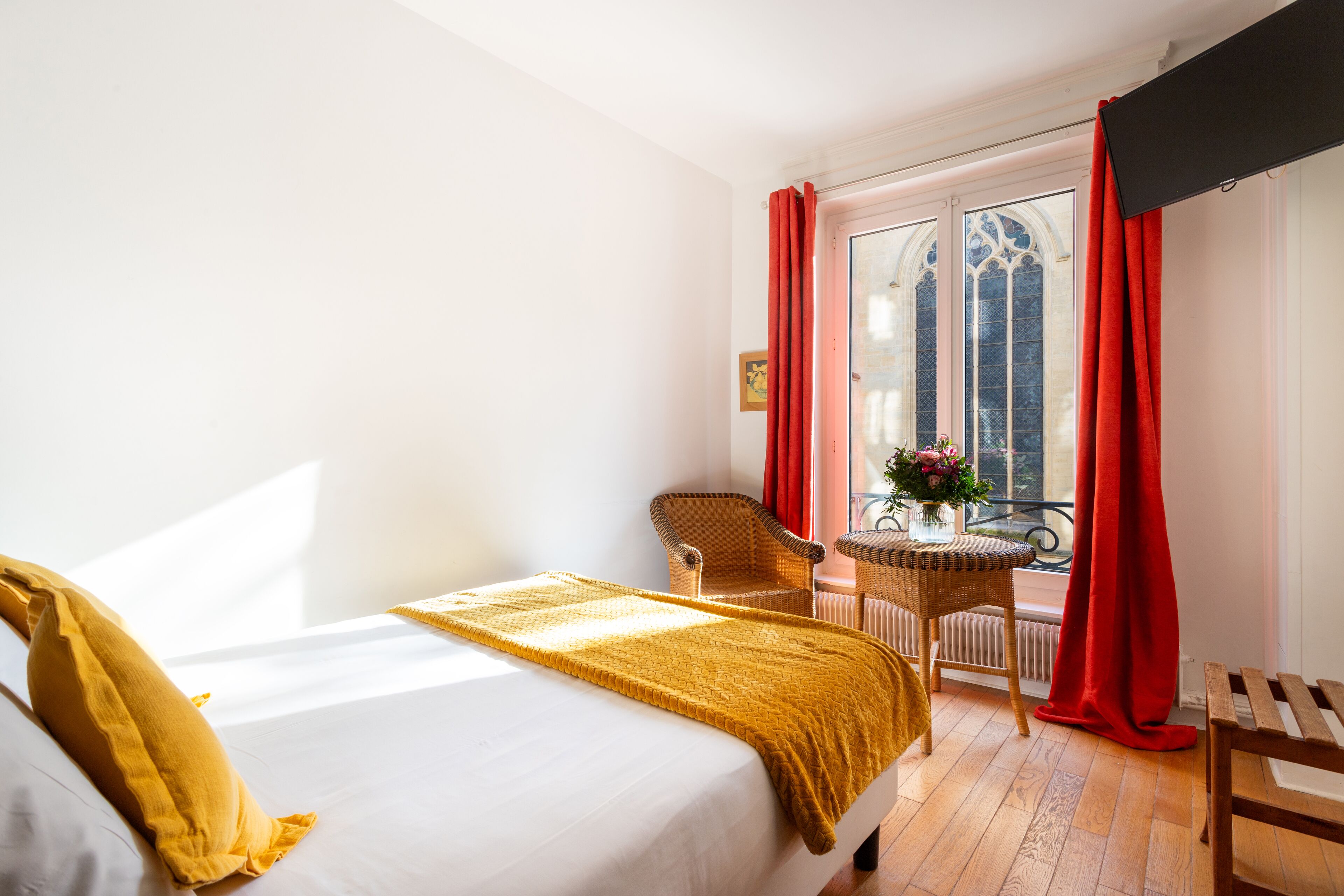 A hotel room with a bed, a chair, a small table with flowers, and a window with red curtains.