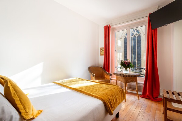 Une chambre d’hôtel avec un lit, une chaise, une petite table ornée de fleurs et une fenêtre avec des rideaux rouges.
