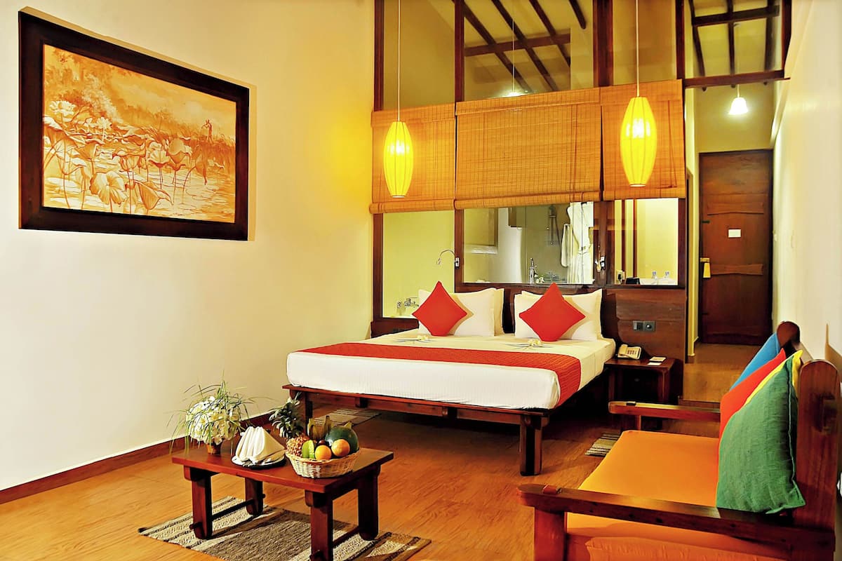 A hotel room with a bed, bedside table with fruit, a chair, a painting on the wall, and a wooden ceiling.